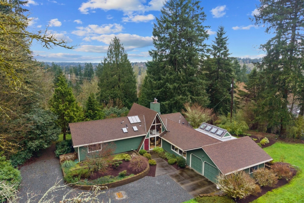 Photo taken of front of home from above showing a green A-frame style house surrounded by acreage, greenery and forest